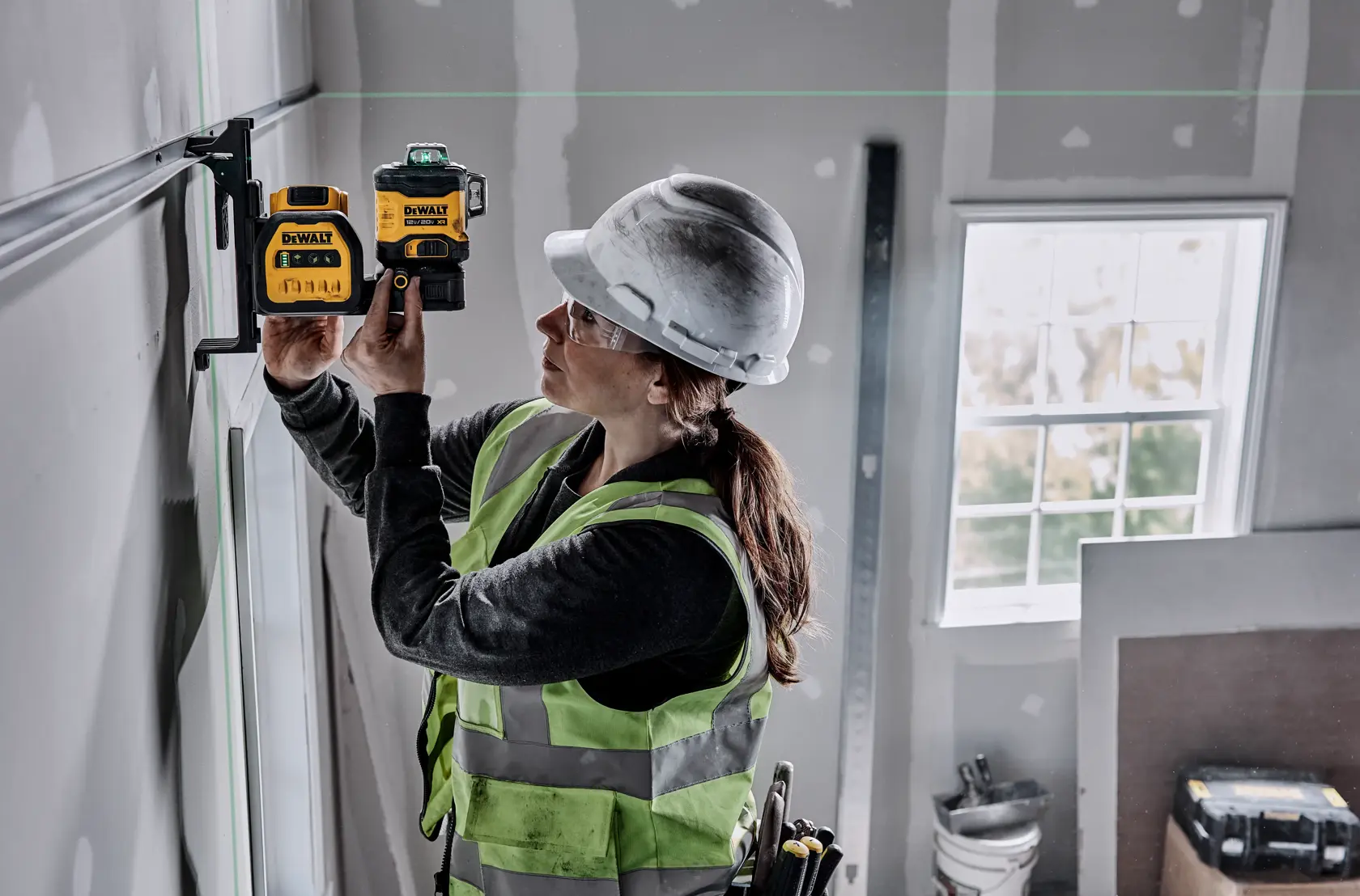 A person wearing a hard hat and safety vest uses a DEWALT green line laser level (model DCLE34030G) to measure or align on a construction site, attaching it to a wall bracket.