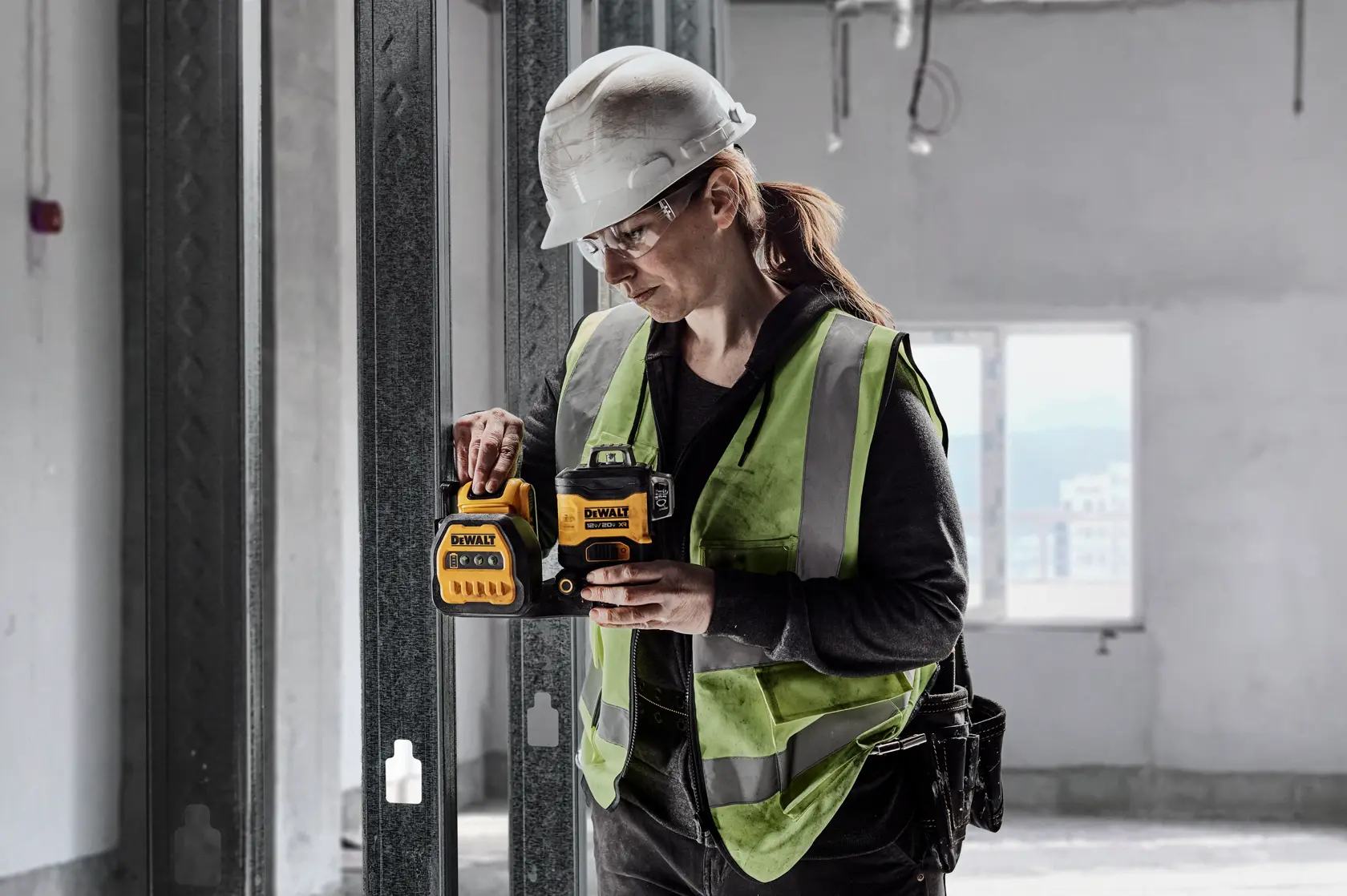 A person wearing a safety helmet and high visibility vest is using a DEWALT DCLE34030G laser level device on a construction site with metal framing and unfinished walls.