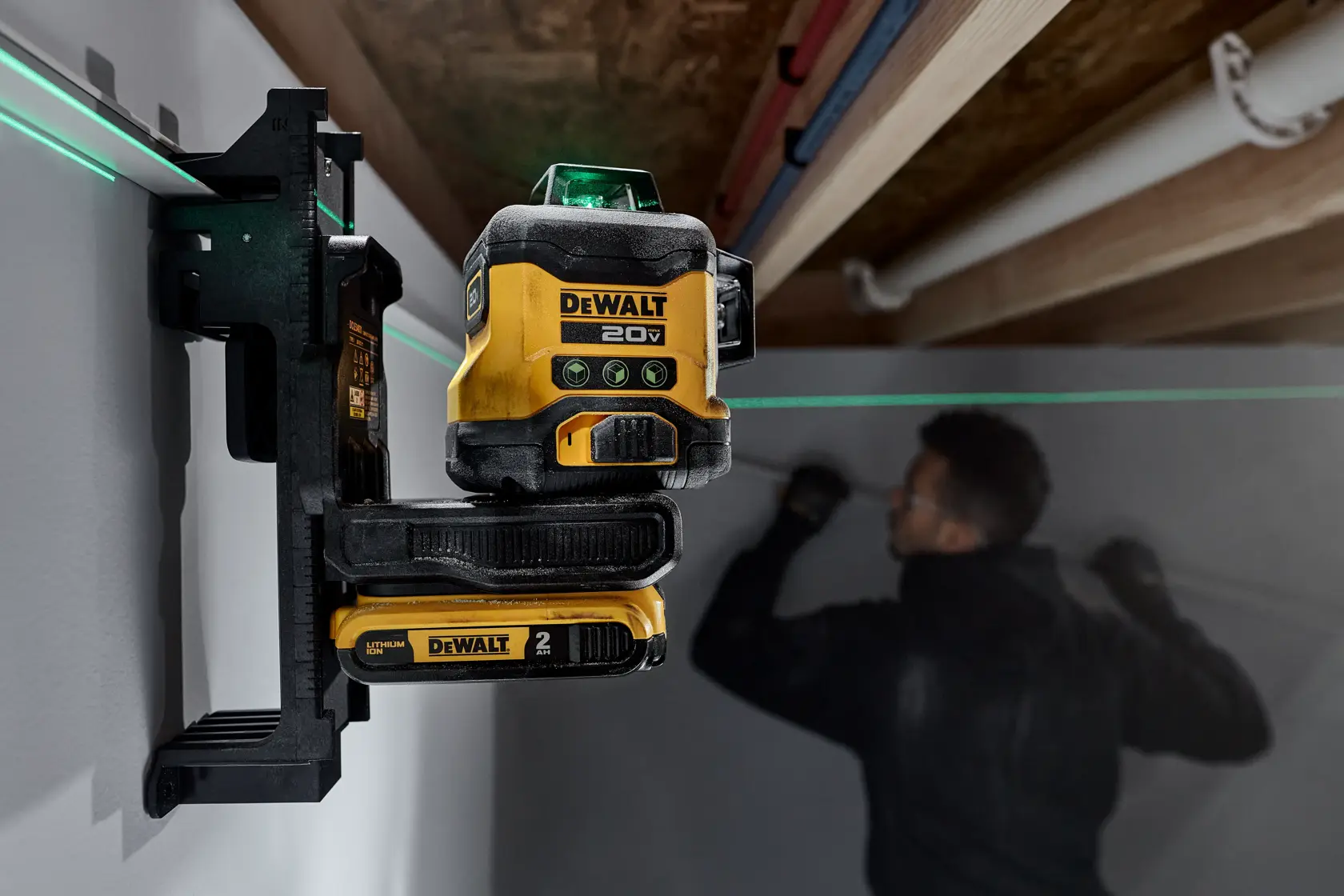 A DEWALT 20V laser level tool mounted on a wall, projecting a green horizontal laser line. A person is in the background working on the wall, with their face blurred.