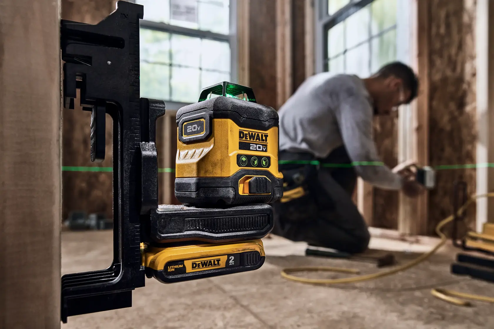 A DEWALT 20V laser level tool mounted on a clamp, projecting a green laser line across a construction site. A person is kneeling in the background, working with tools on wooden framing.