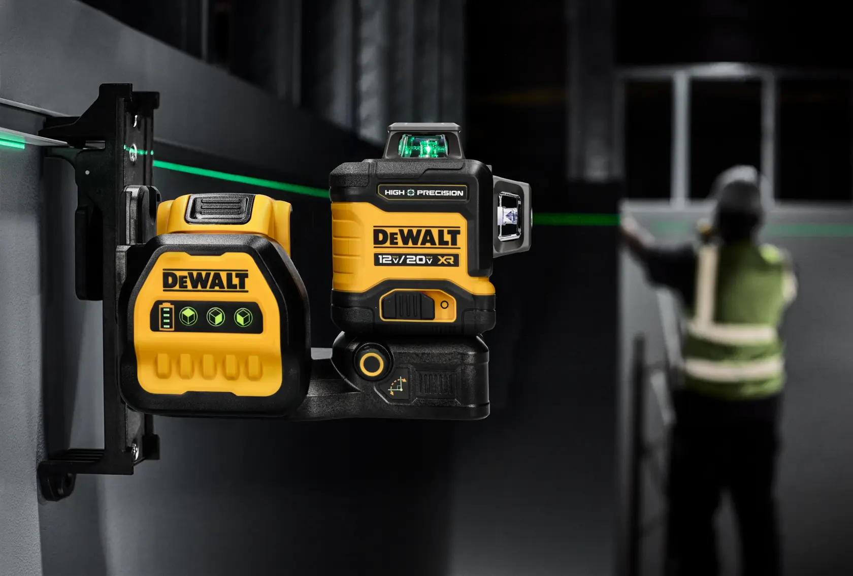 A DEWALT 12V/20V XR high precision green laser level is mounted on a wall bracket. It emits a visible green laser line across the workspace. The device is yellow and black, featuring DEWALT branding. In the background, a person wearing a reflective vest is working near the laser line.
