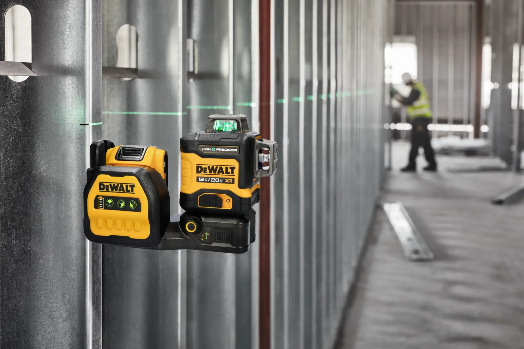 A DEWALT laser level device is mounted on a metal construction wall, projecting a green horizontal laser line across the room. In the background, a person wearing a safety vest is working in the unfinished industrial space.