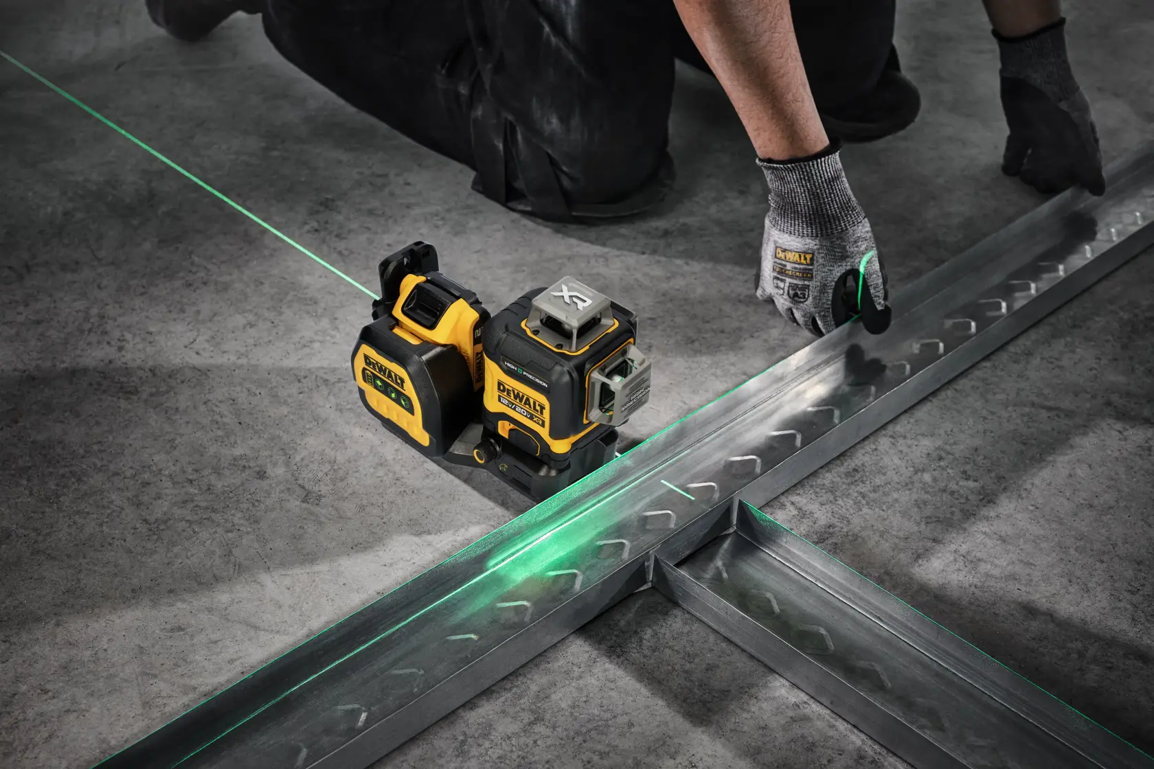 A DEWALT laser level device is mounted on a metal construction wall, projecting a green horizontal laser line across the room. In the background, a person wearing a safety vest is working in the unfinished industrial space.