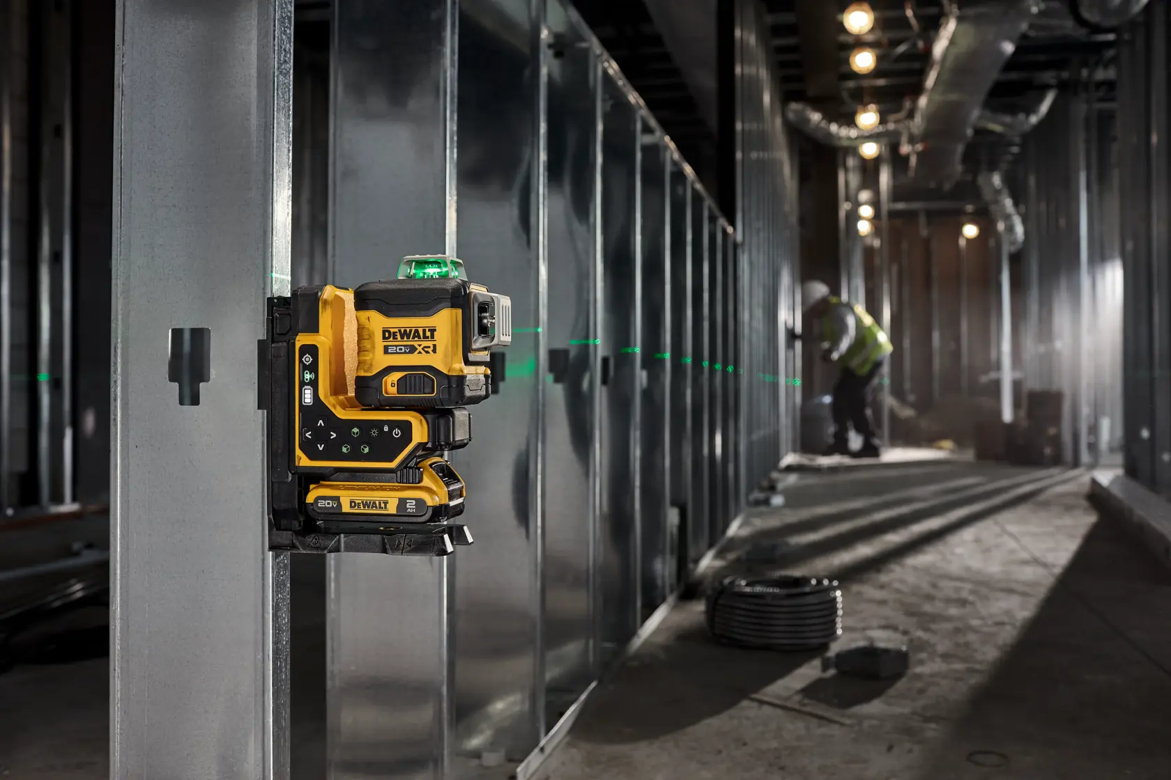 DEWALT laser level mounted on metal wall, emitting green line in construction area with worker in background.