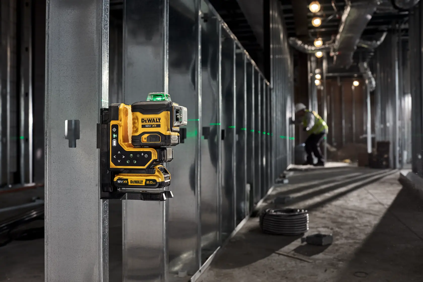 DEWALT laser level mounted on metal framing, projecting green lines; worker in background of construction site.