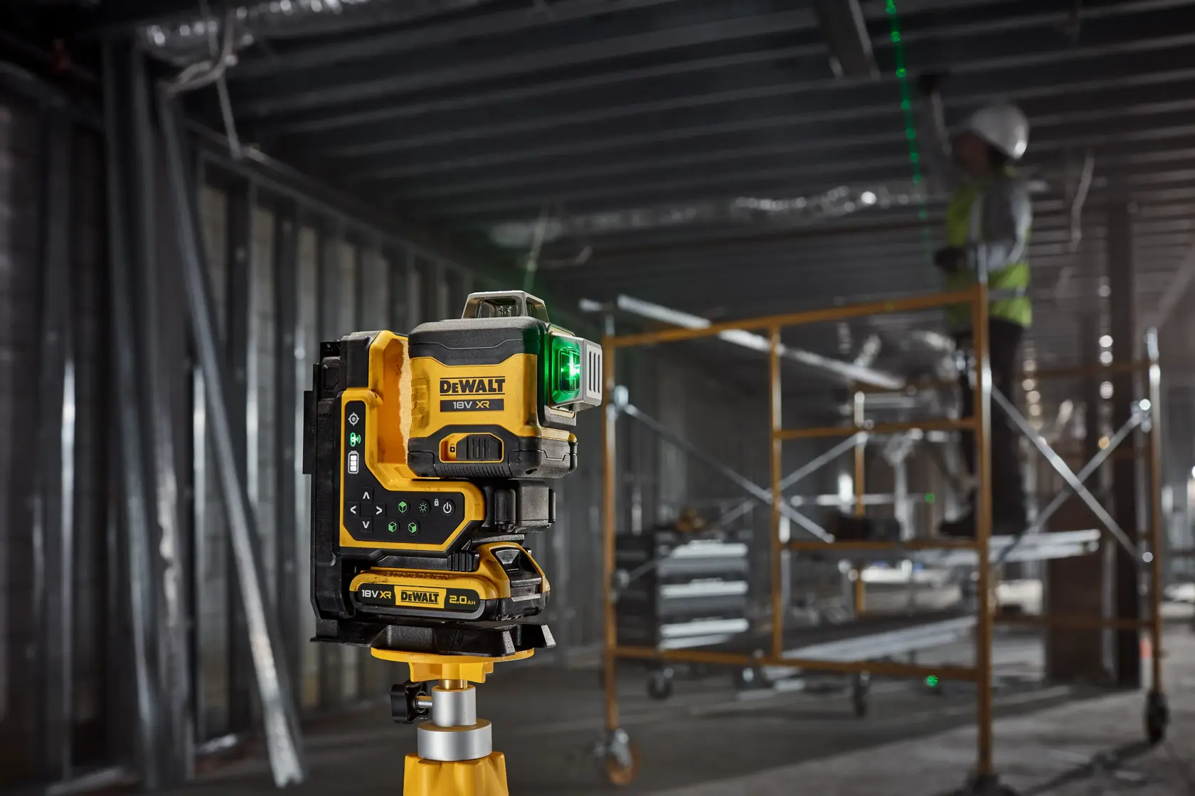 DEWALT laser level with green beam on tripod at construction site, worker and scaffolding in background.