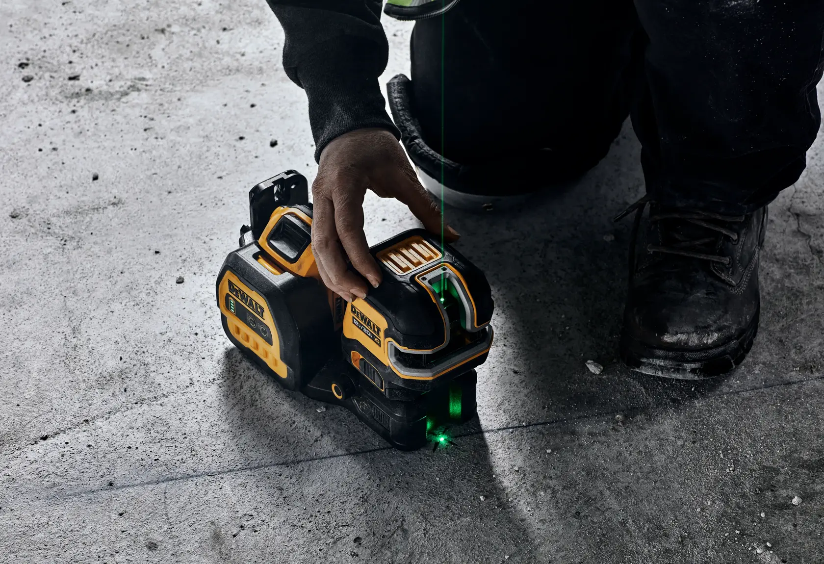 A close-up of a person using a DEWALT green line laser level tool on a concrete floor. The tool projects a green laser line across the surface for alignment purposes.