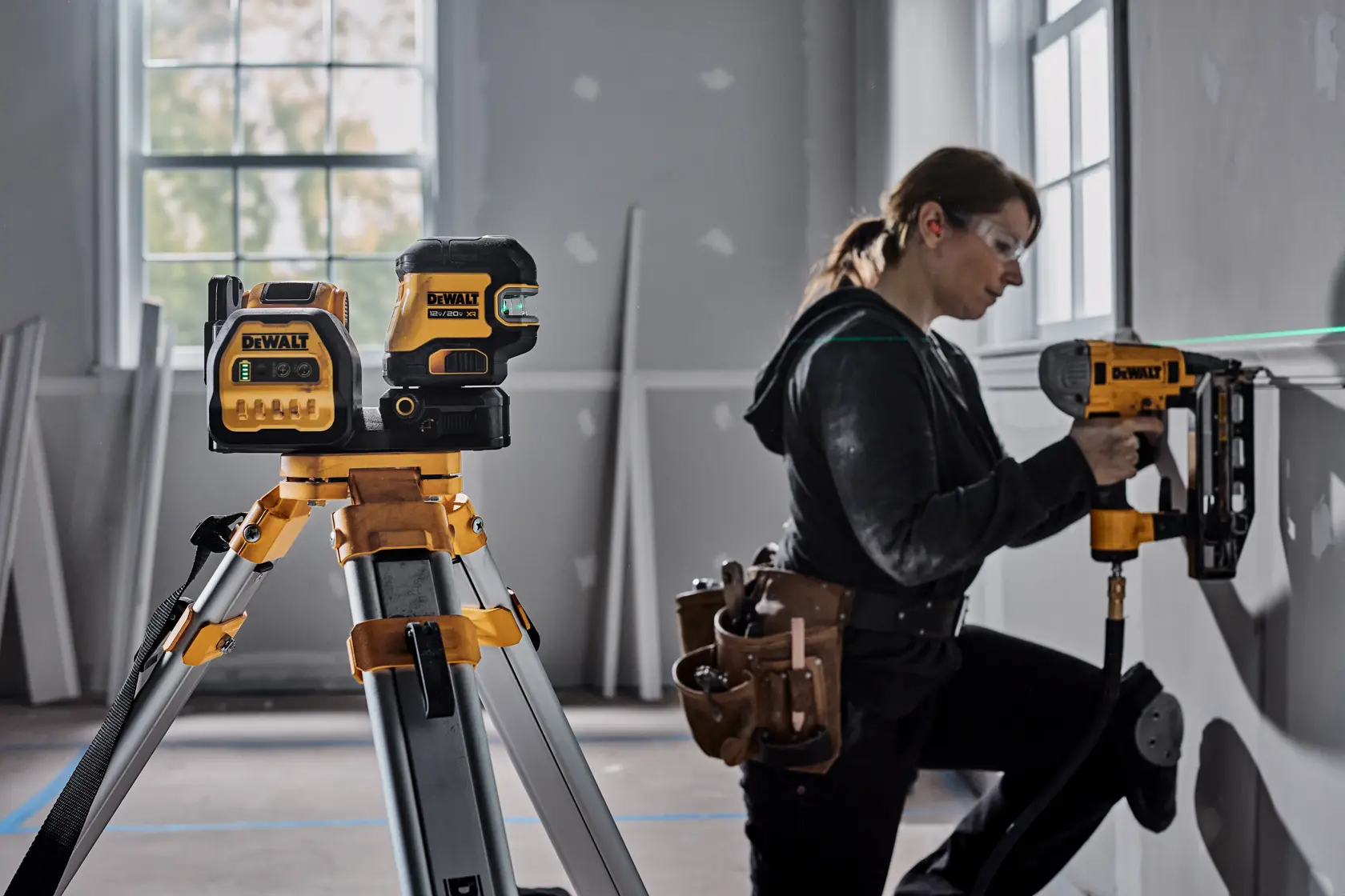 A DEWALT laser level tool is mounted on a tripod in the foreground of a room under construction. In the background, a person with their face blurred is using a DEWALT power tool to install material on a wall. Various construction materials and tools can be seen around the room.