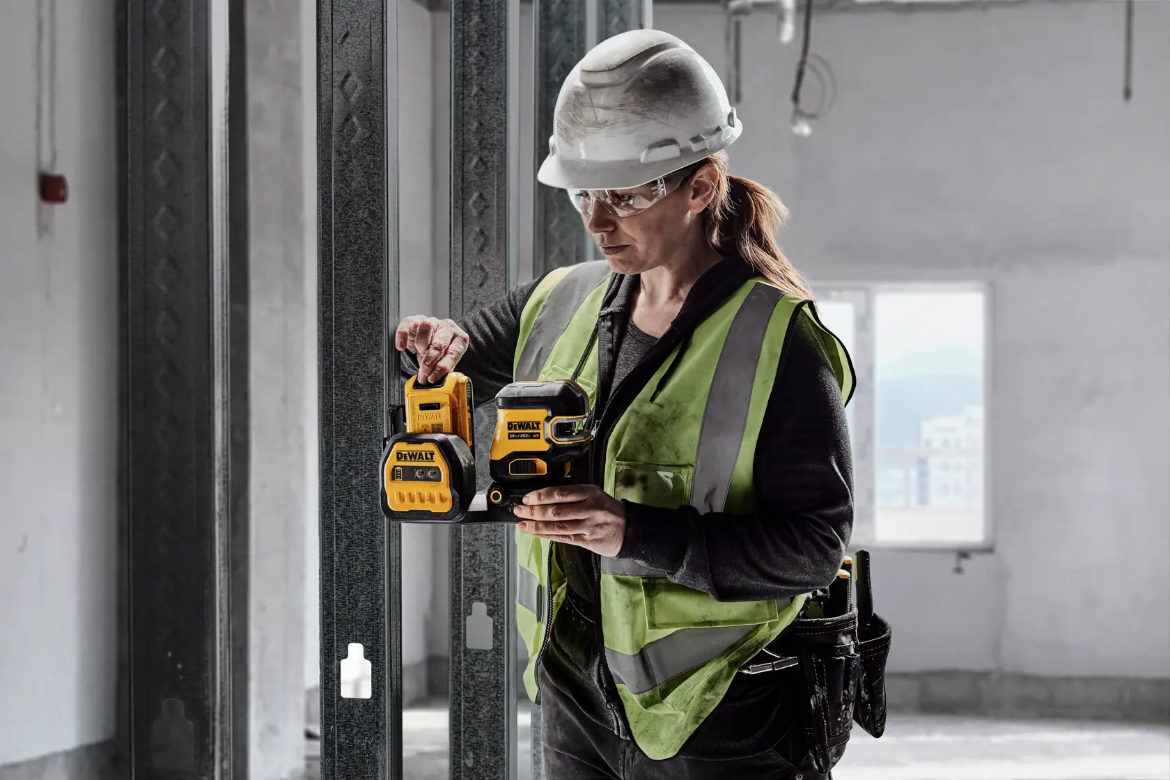 A person wearing a safety helmet and high-visibility vest is holding a DEWALT DCLE34220G green cross line and laser level in a construction environment.