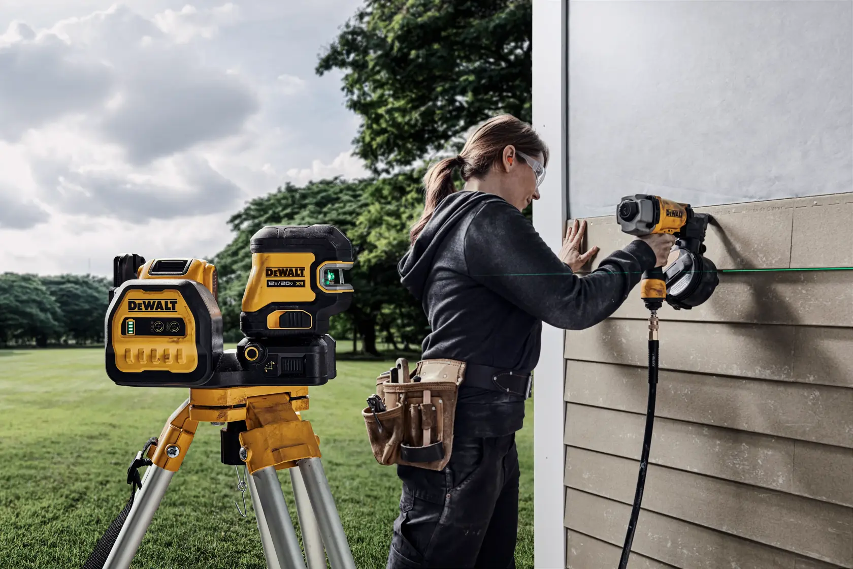 A person using DEWALT tools, including a DEWALT laser level on a tripod, to install siding panels on an exterior wall. The person is wearing a tool belt and working outdoors.