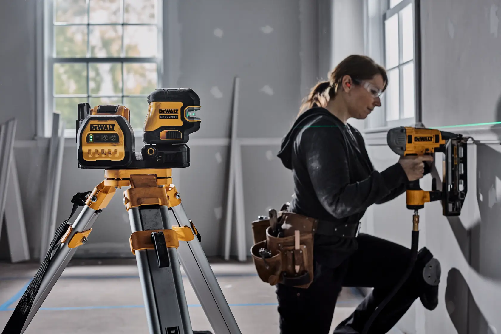 A DEWALT green laser level is mounted on a tripod in a room under construction. A person with a tool belt is using a DEWALT nail gun on a wall, aligning their work with the green laser line. Various construction tools are visible in the room.