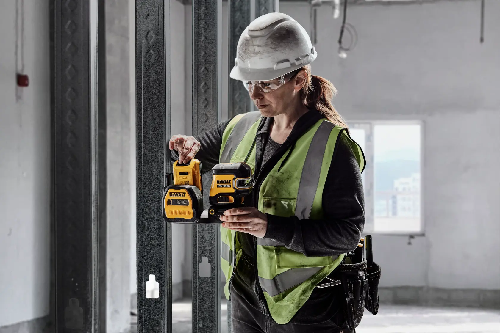A construction worker wearing a green safety vest and white hard hat is holding and operating a DEWALT laser level tool inside a building under construction.