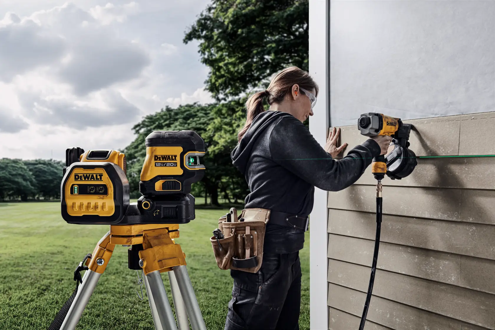 A DEWALT laser level on a tripod is positioned outdoors, aimed at a wall where a person wearing a tool belt is using a nail gun to install siding. The scene is set in a grassy area with trees in the background.