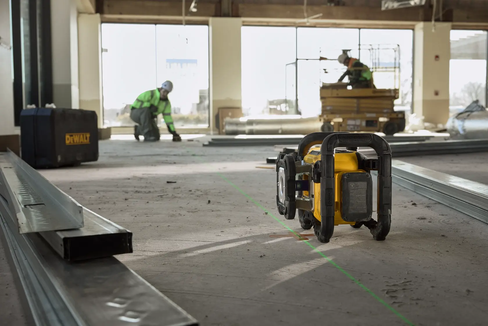 Un outil de niveau laser DEWALT est placé sur le sol dans un site de construction, projetant une ligne de faisceau vert. Des montants métalliques et une valise DEWALT sont visibles, avec deux ouvriers en équipement de sécurité travaillant en arrière-plan.