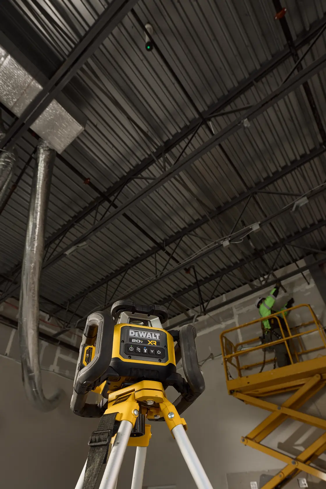 Un niveau laser DEWALT 20V XR est monté sur un trépied dans un environnement industriel, avec un travailleur sur une nacelle jaune en arrière-plan travaillant près du plafond.