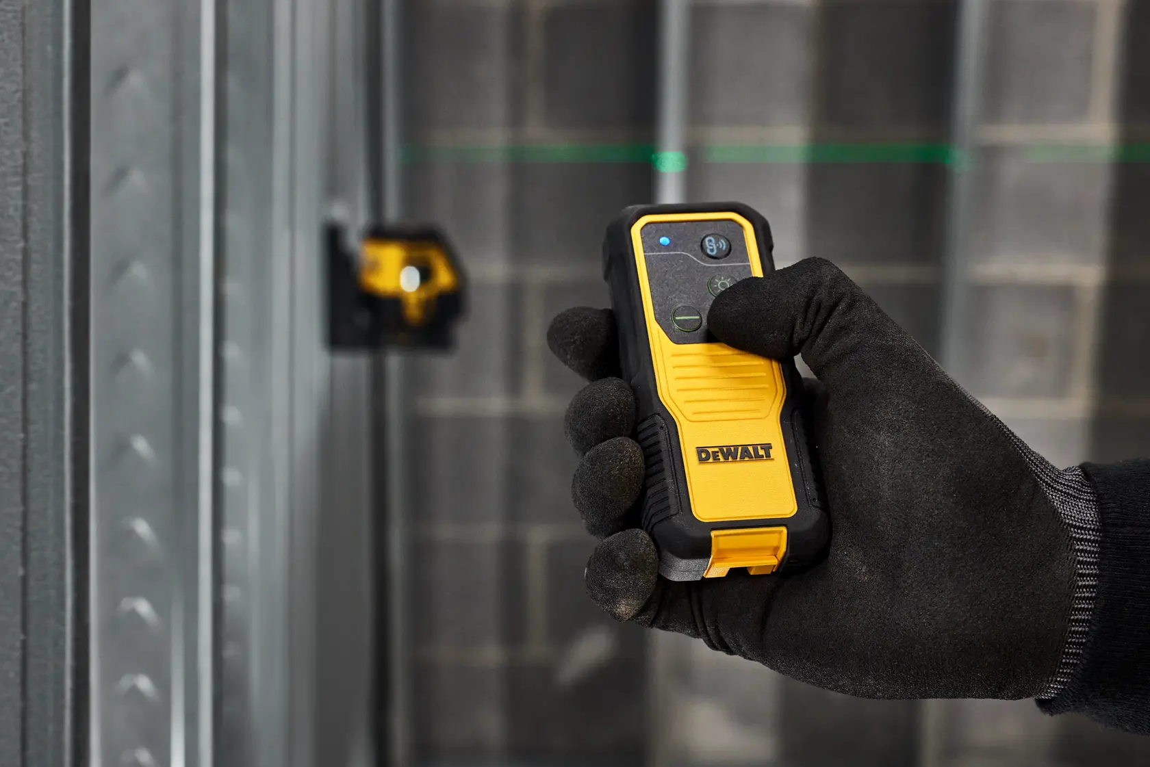 A gloved hand holding a DEWALT laser detector device, with a DEWALT laser level projecting green laser lines onto a wall in the background. The setting appears to be a construction site with metal framing and concrete blocks.