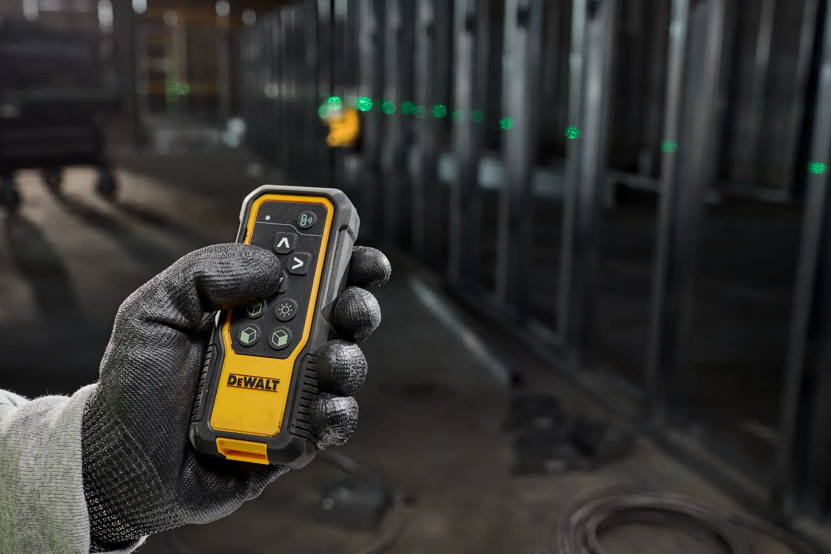 A gloved hand holds a DEWALT remote control device in an industrial setting. In the background, a green laser line is visible, used for alignment during construction.