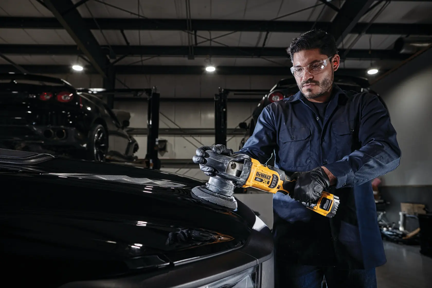 A person wearing safety glasses and gloves uses a DEWALT DCM848P2 cordless polisher to buff the hood of a black car in a garage with other vehicles in the background.