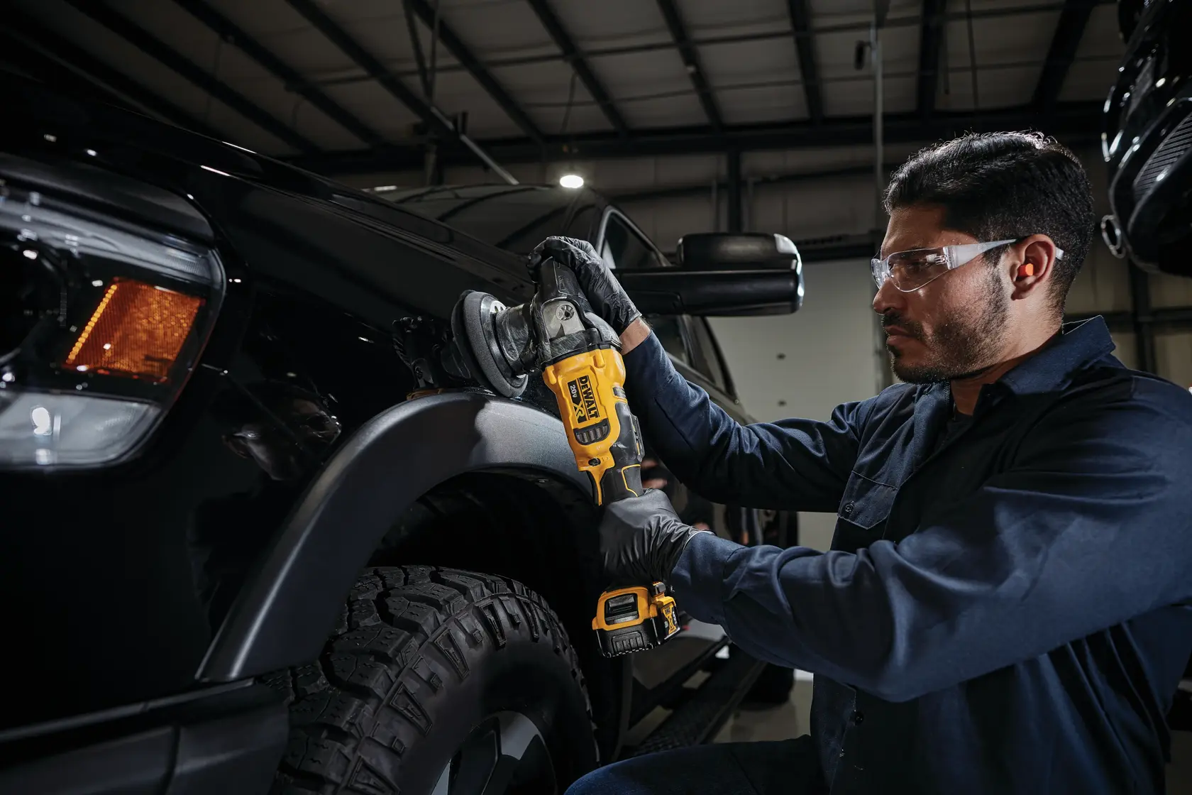 A person wearing black gloves uses a DEWALT cordless polisher to buff the surface of a black vehicle inside a garage. The person's face is blurred for privacy.