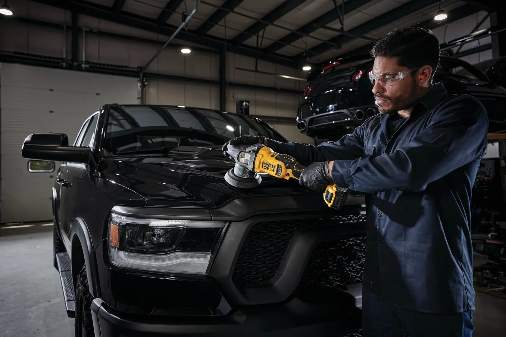 A person using a DEWALT cordless polisher to polish the hood of a black pickup truck inside a garage. The face of the person is blurred, and they are wearing dark clothing and gloves.