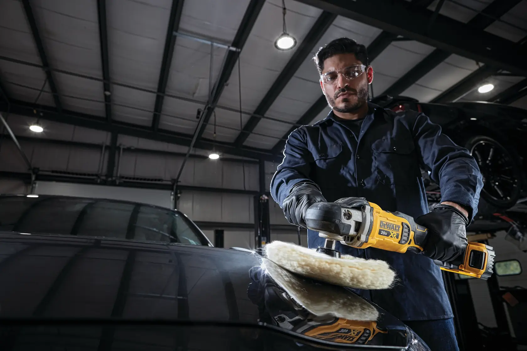 A person wearing gloves and a dark work uniform is polishing the hood of a car with a yellow DEWALT cordless polisher inside an industrial garage.