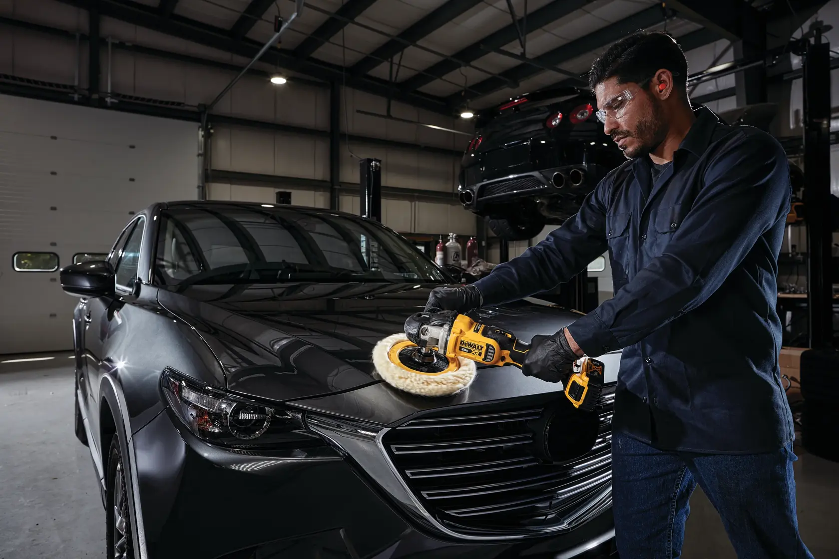 A person wearing gloves uses a DEWALT cordless polisher (model DCM849P2) to polish the hood of a dark-colored car inside an automotive workshop.