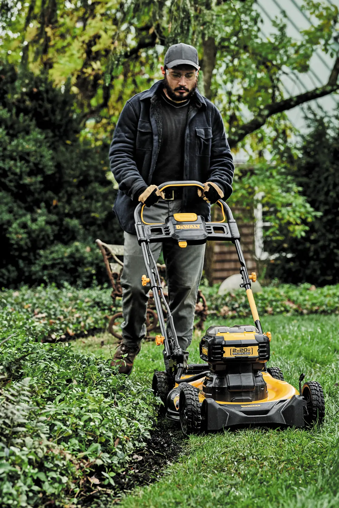 A person is using a DEWALT battery-powered lawn mower on a grassy lawn with shrubs nearby. The person’s face is blurred and they are wearing a jacket and gloves. The mower is black and yellow, and the scene is outdoors with trees in the background.