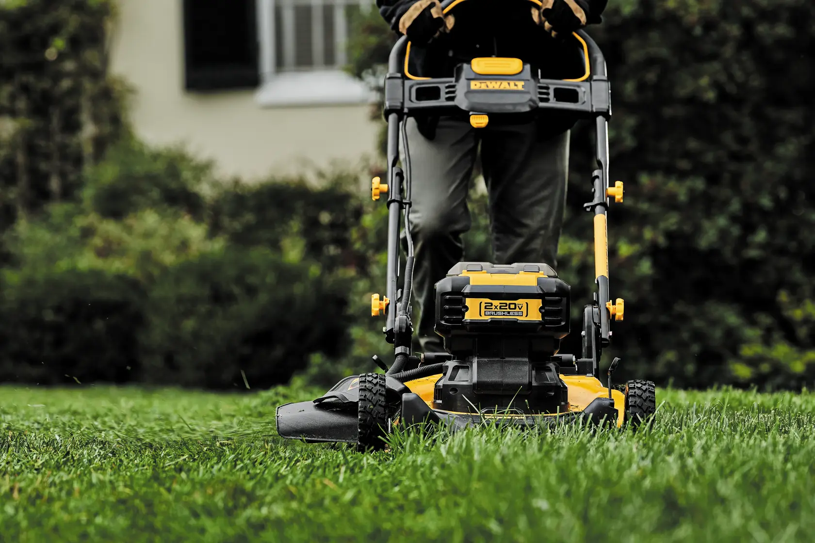 A person is pushing a yellow and black DEWALT cordless lawn mower across a grassy lawn, with the mower's brand and battery compartment visible.