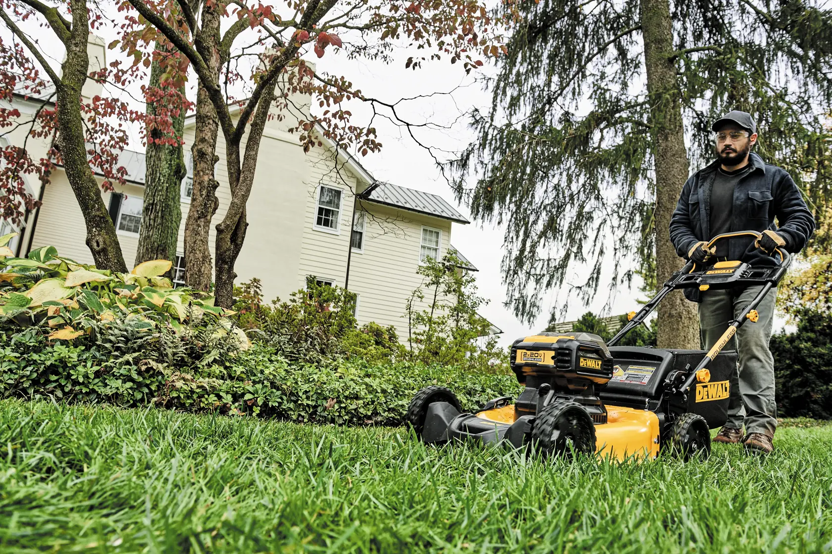 A person mowing the lawn with a yellow DEWALT cordless lawn mower, surrounded by green grass, plants, and trees, with a white house visible in the background.