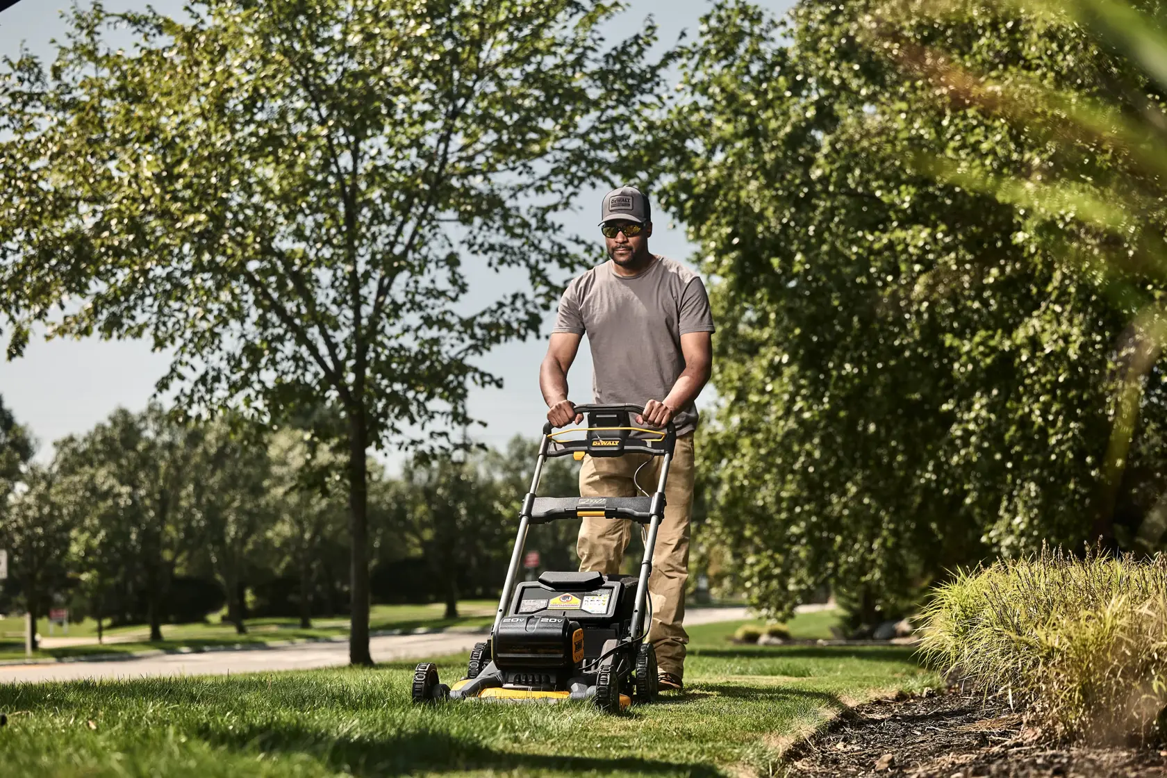 A person is mowing the lawn with a DEWALT cordless lawn mower outdoors on a sunny day. Green grass and leafy trees surround the area.