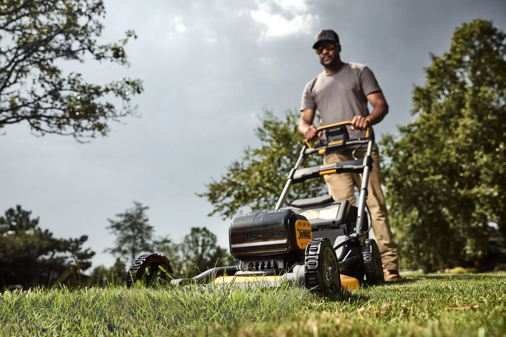 A person using a DEWALT cordless lawn mower to cut grass in a yard. The focus is on the mower, with trees and a cloudy sky in the background.