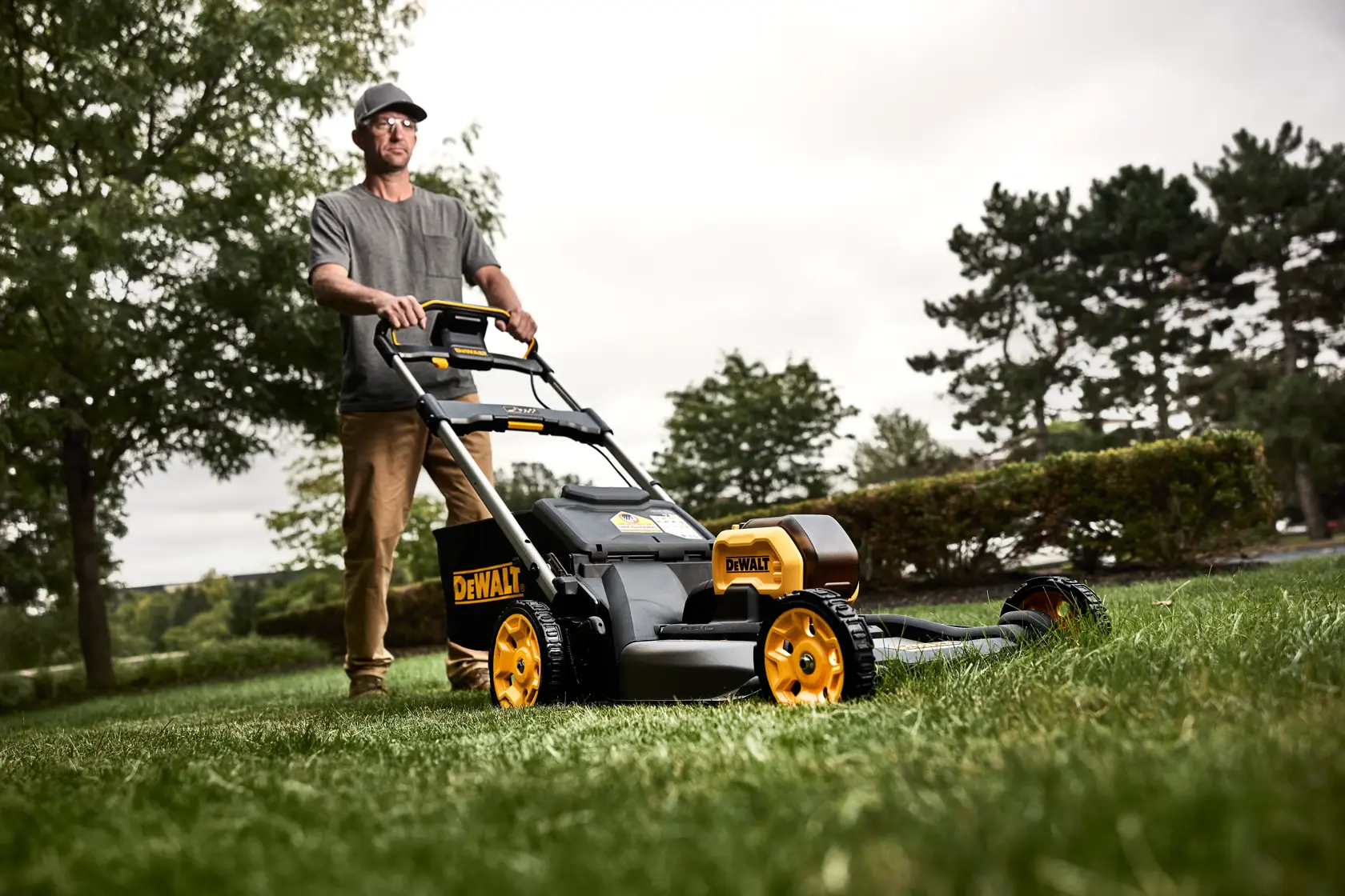 A person using a DEWALT lawn mower outdoors on a grassy lawn. The lawn mower is black and yellow and appears to be battery-powered. Trees and bushes are visible in the background.