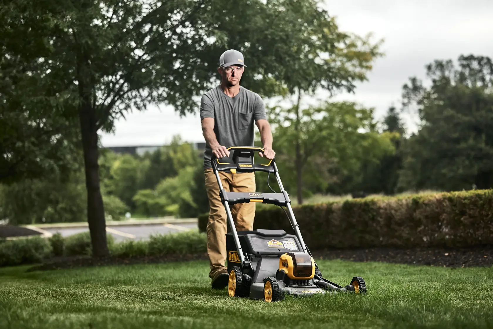 A person operating a DEWALT lawn mower model DCMWP600X2 on a grassy lawn outdoors. The lawn mower is black and yellow with visible branding, and the scene includes trees and bushes in the background.