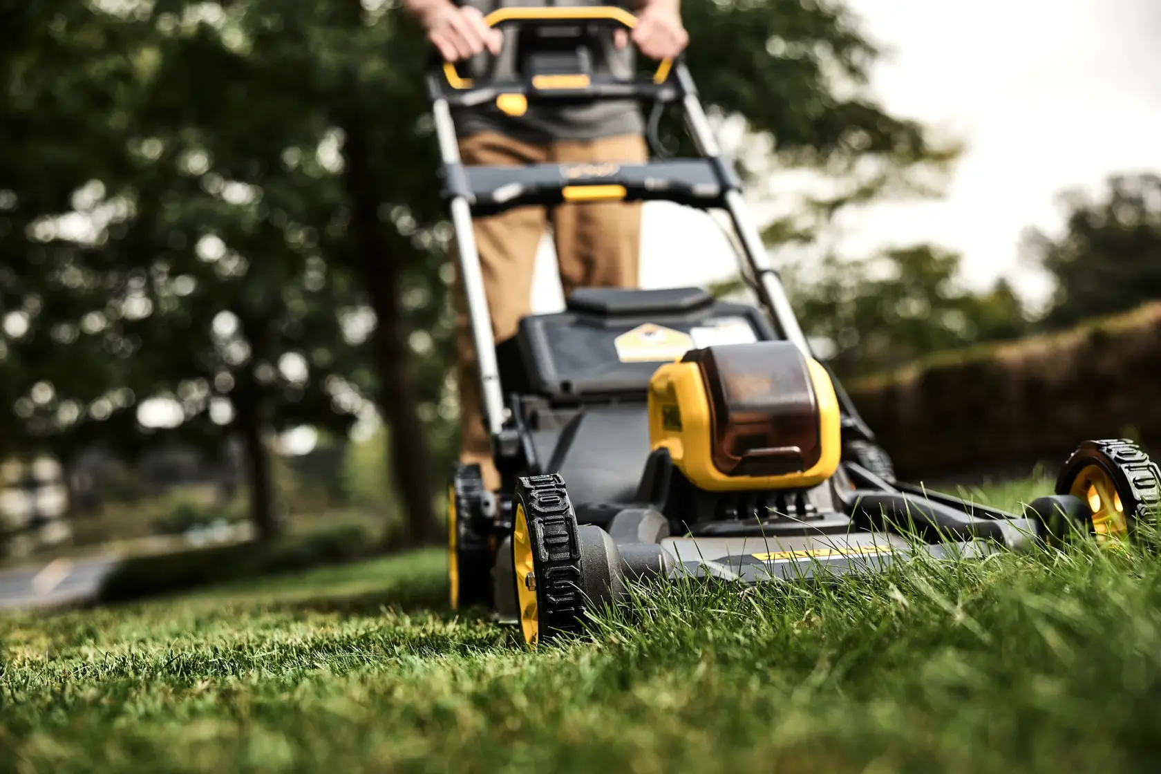 A close-up view of a DEWALT cordless lawn mower being used outdoors on grass, with the operator's hands visible on the handle. The mower features yellow and black accents.