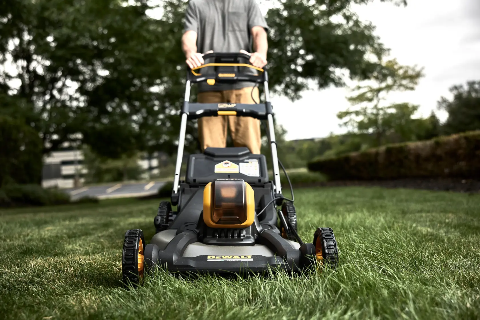 A DEWALT lawn mower is being pushed on grass outdoors by a person. The mower is black and yellow, and the person is visible from the waist down. Trees and shrubbery are in the background.