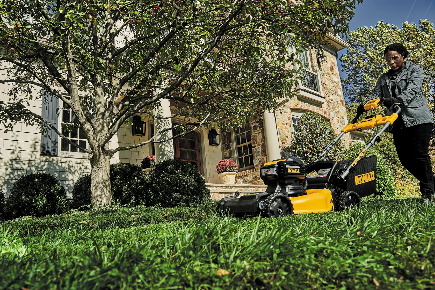 A person is pushing a DEWALT lawn mower across a green lawn in front of a house with stone and siding walls, trees, and shrubs.