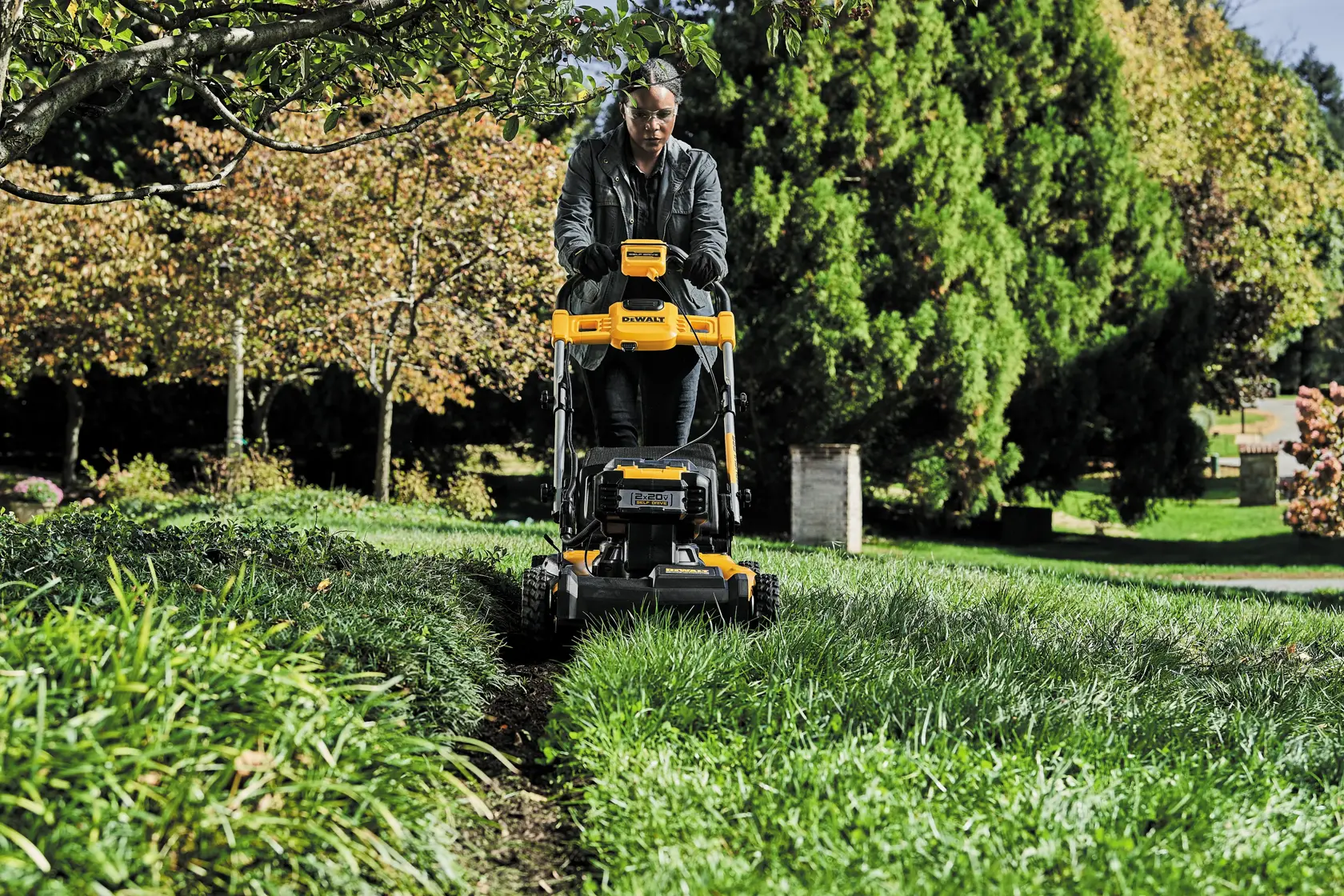 A person mowing the lawn with a DEWALT cordless lawn mower in a garden with green grass, trees, and autumn foliage.
