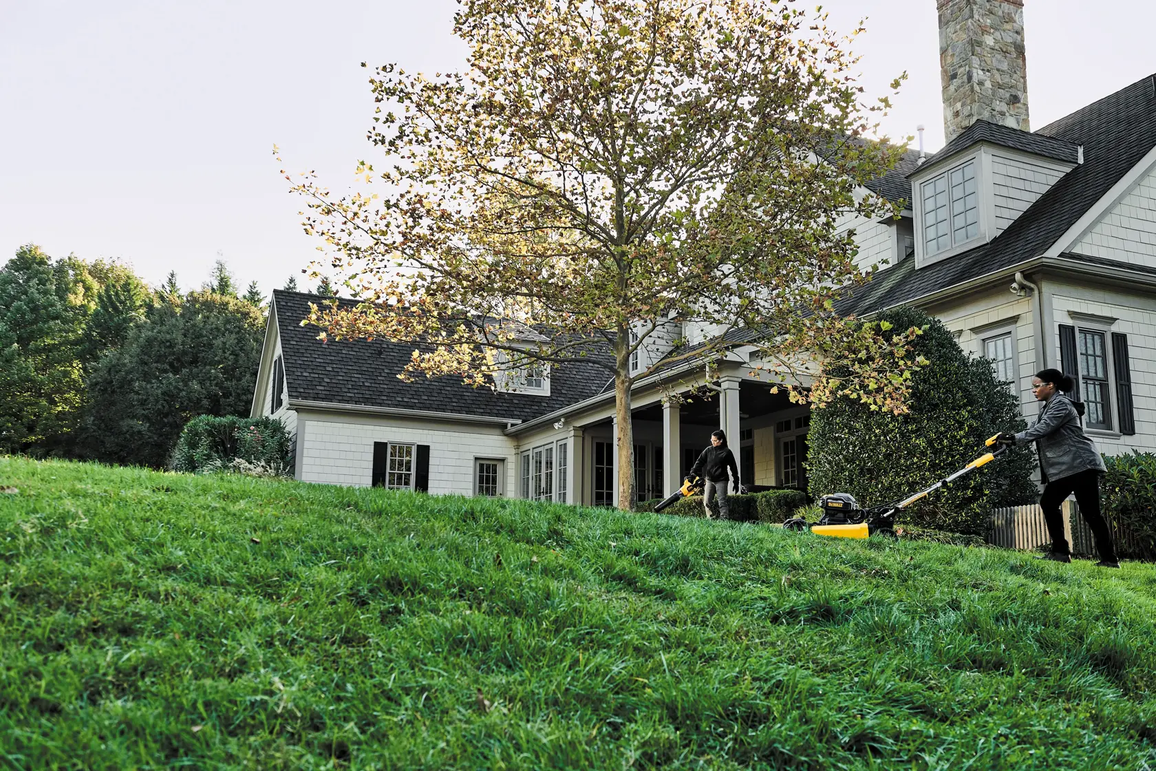 Two people are using yellow and black lawn mowers to cut grass on the lawn in front of a large house with a porch and a tree.