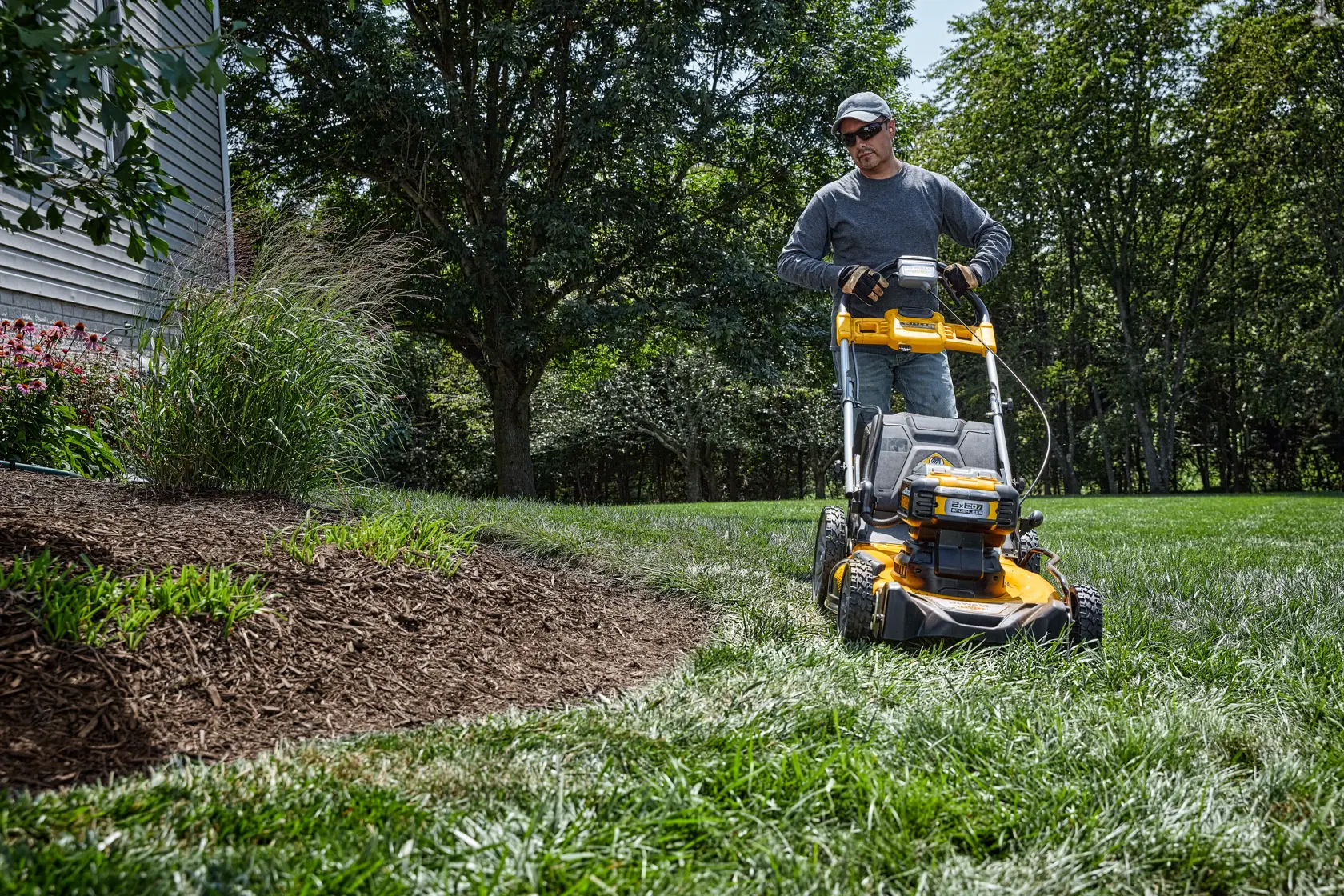 A person is using a yellow DEWALT lawn mower to cut grass near a landscaped garden bed with mulch and plants. The scene is outdoors with trees and greenery in the background.
