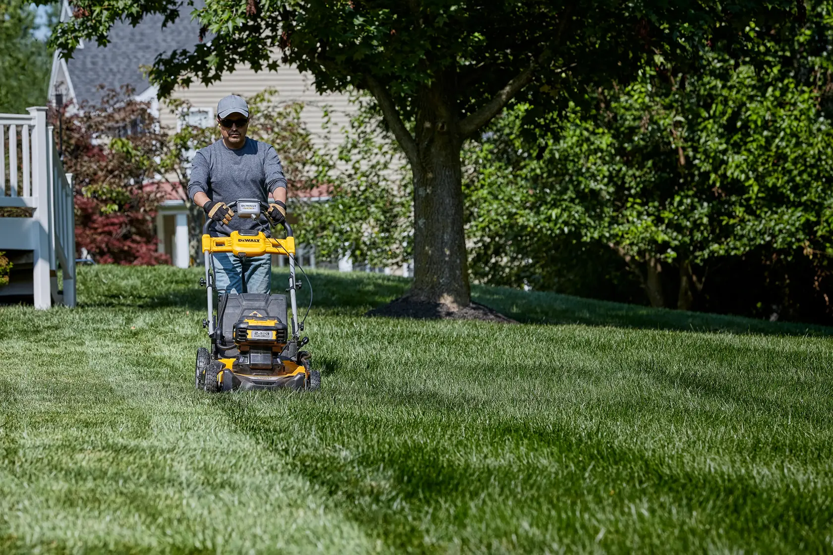 A person is using a yellow DEWALT lawn mower to cut grass in a residential yard. The lawn mower is being pushed across a green lawn, with trees and a house in the background.