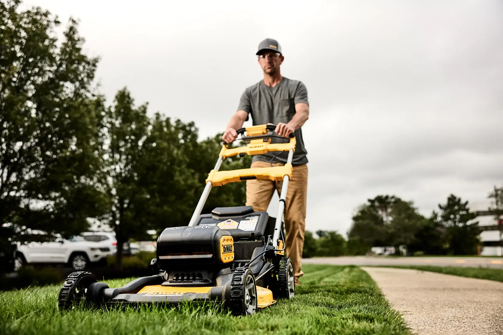 A person is pushing a DEWALT lawn mower on grass beside a walkway, with trees and cars visible in the background. The person's face is blurred and they are wearing a grey shirt and khaki pants.