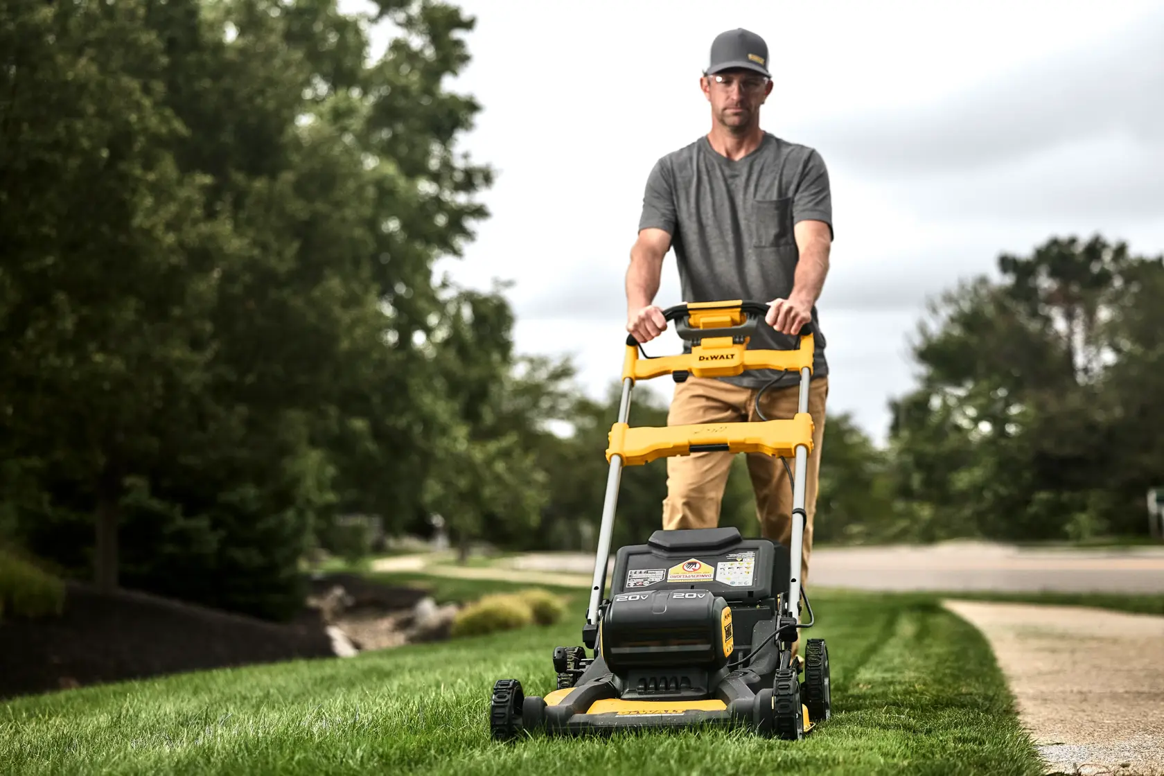 A person is pushing a yellow and black DEWALT lawn mower on a grassy lawn beside a sidewalk, with trees and a road in the background.