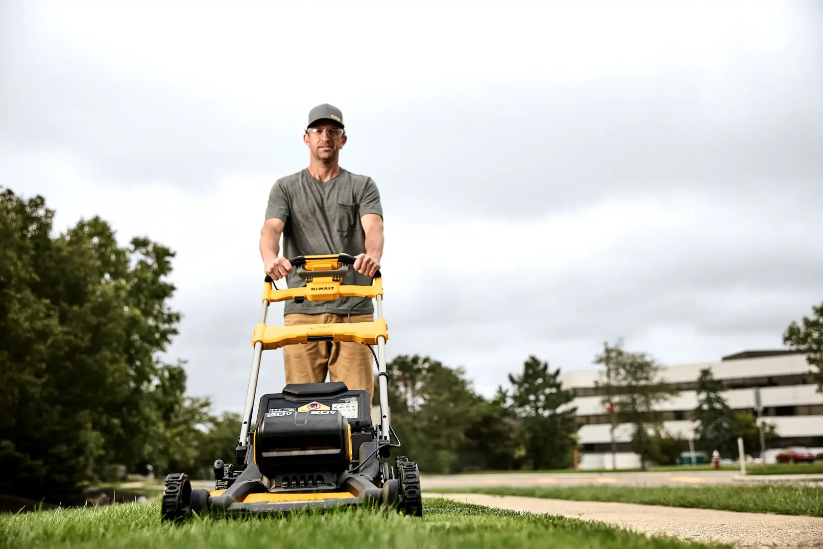A person is pushing a DEWALT lawn mower model DCMWSP256U2 on a grassy area outdoors. The lawn mower is predominantly yellow and black, and there are buildings and trees in the background.