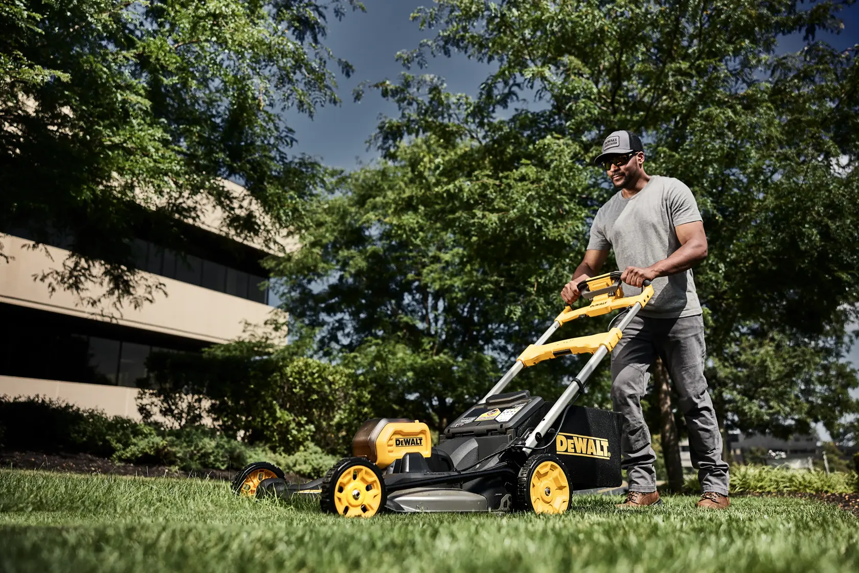 A person is pushing a DEWALT DCMWSP650Y2 lawn mower on green grass in a landscaped outdoor area, with trees and a building in the background.