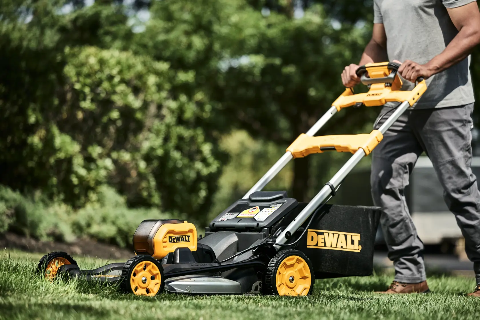 A person is using a DEWALT lawn mower with yellow and black accents to cut grass in an outdoor garden. The mower features a grass collection bag and battery compartment. Trees and plants can be seen in the background.
