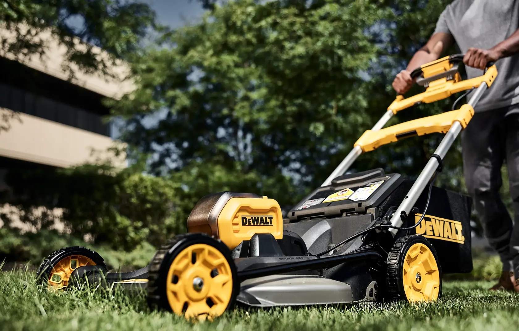 A person pushes a DEWALT cordless electric lawn mower with yellow and black detailing across a green lawn. The mower is shown in use outdoors, with trees and a building visible in the background.