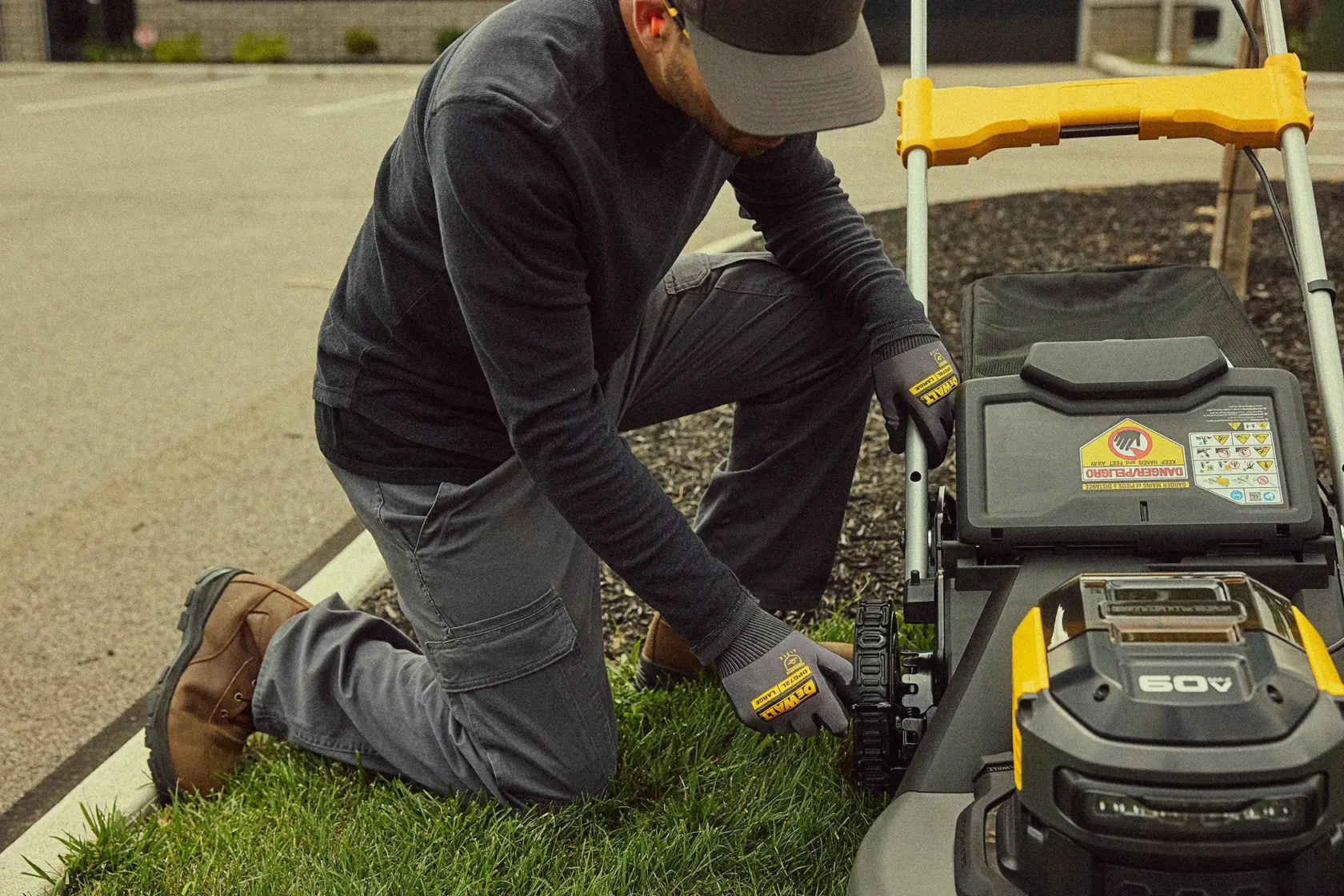 A person wearing gloves kneels on grass and adjusts a DEWALT 60V lawn mower near a curbside. The person's face is blurred and they are dressed in work boots and cargo pants.