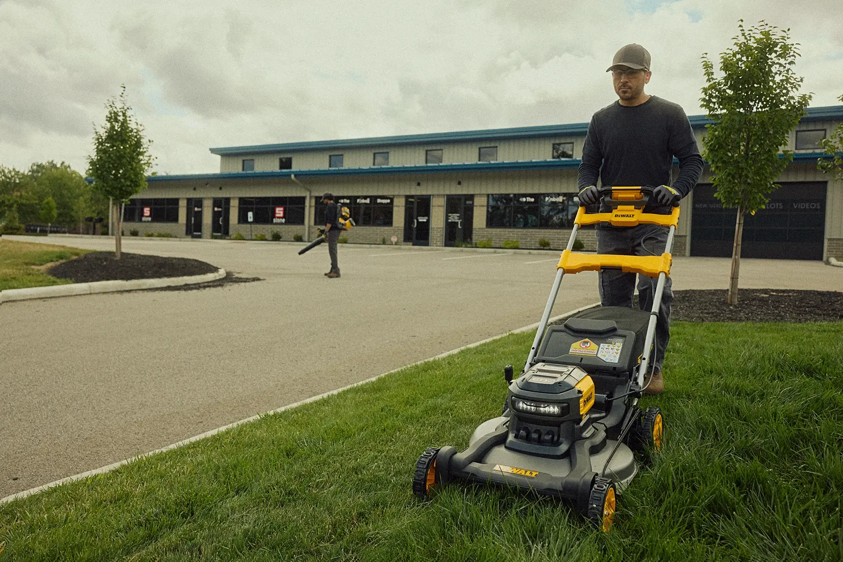 A person is using a DEWALT lawn mower to cut grass near a commercial building. Another person in the background is using a leaf blower. The setting appears to be a landscaped area with trees and grass.