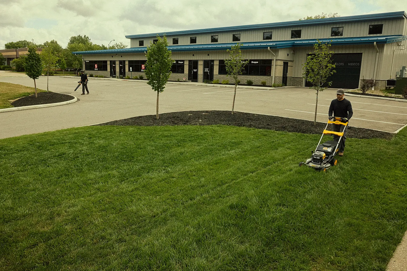 A person using a DEWALT lawn mower to cut grass in front of a commercial building, with another person in the background working with equipment near the parking lot.