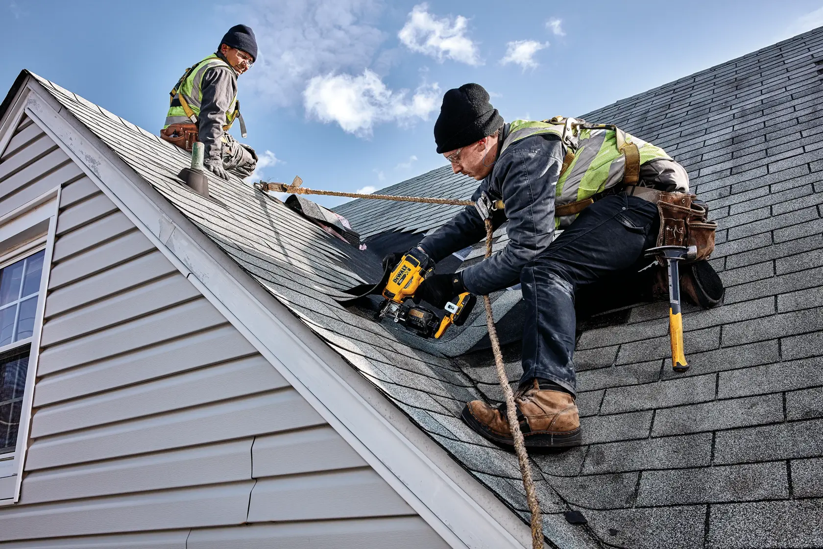 Two workers in safety gear are installing shingles on a house roof. One worker is using a yellow DEWALT cordless roofing nailer, while the other is seated further up the roof. The workers are secured with ropes and appear to be performing roofing tasks under a bright blue sky.